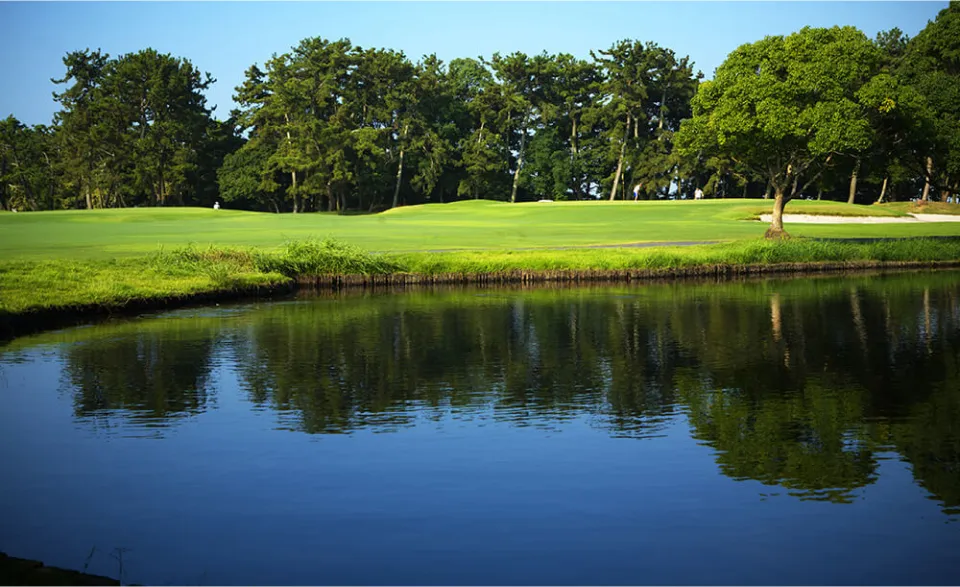 A serene golf course with a calm lake in the foreground, reflecting trees and greenery from the course under a clear blue sky.