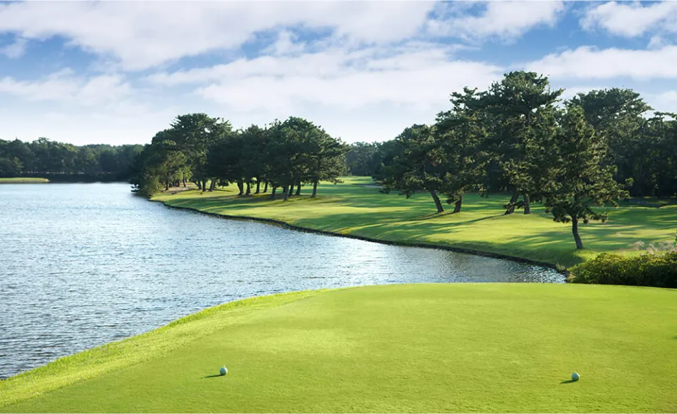 A scenic golf course with a lake on the left and a well-maintained green on the right, surrounded by trees under a bright sky.