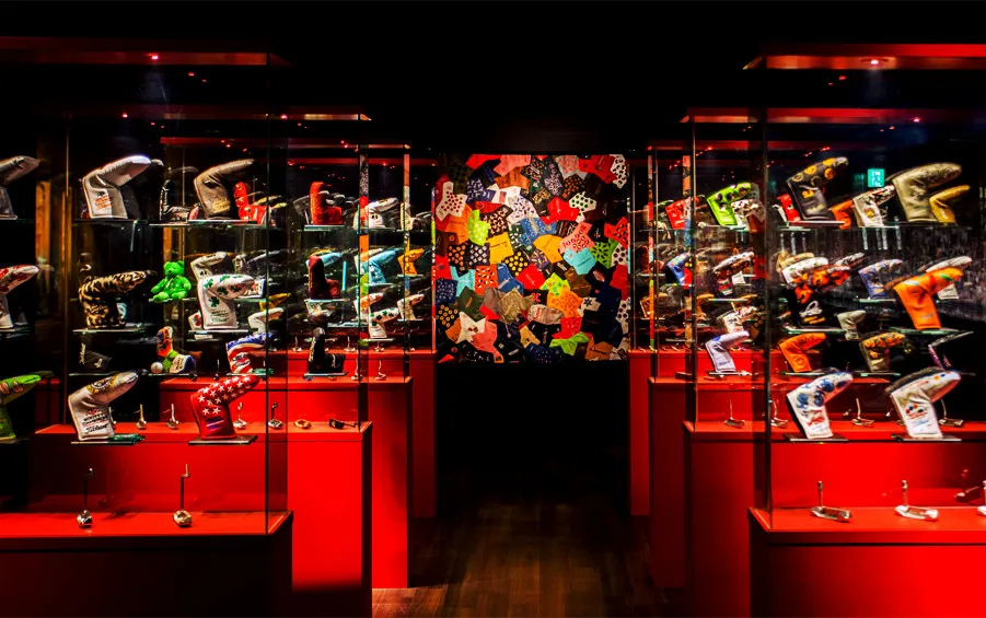 A vibrant exhibit of golf putter covers in various designs, displayed in glass cases in a red-lit gallery.